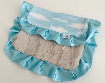 May include: A baby blanket with a light blue satin border and a soft, plush center. The top layer features a sky blue fabric with white cloud patterns. The blanket has a tag that reads "Brown Dane Designs".
