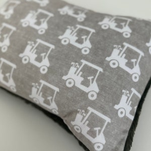 May include: A rectangular gray pillow with a repeating pattern of white golf carts. The pillow has a dark gray, fuzzy border. The golf carts are detailed with wheels, steering wheels, and golf club sets.