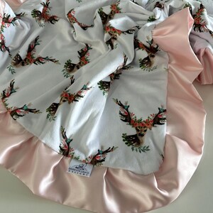 May include: A white baby blanket with a pink satin trim. The blanket features a repeating pattern of deer heads with floral wreaths. The blanket has a tag that reads "Southern Grace Designs".