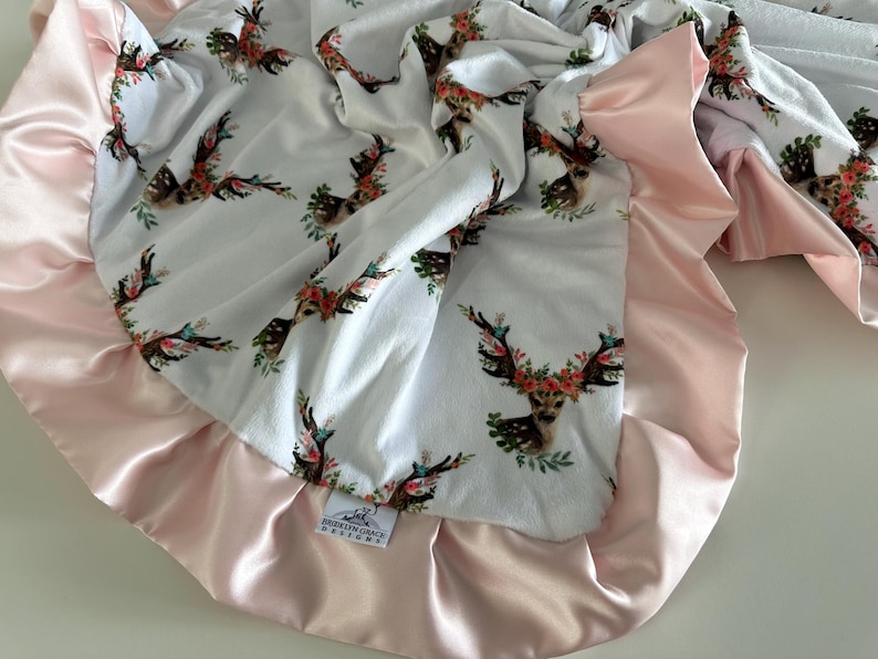 May include: A white baby blanket with a pink satin trim. The blanket features a repeating pattern of deer heads with floral wreaths. The blanket has a tag that reads "Southern Grace Designs".