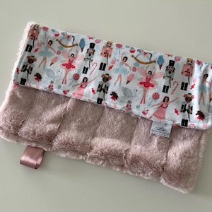 May include: A pink plush baby blanket with a white cotton fabric on one side featuring a Nutcracker and ballerina print. The blanket has a satin ribbon attached.