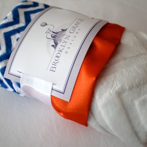 May include: A white and blue chevron patterned baby blanket with a white and orange satin ribbon. The blanket is folded and tied with the ribbon. The blanket is labeled "Brooklyn Grace Designs".