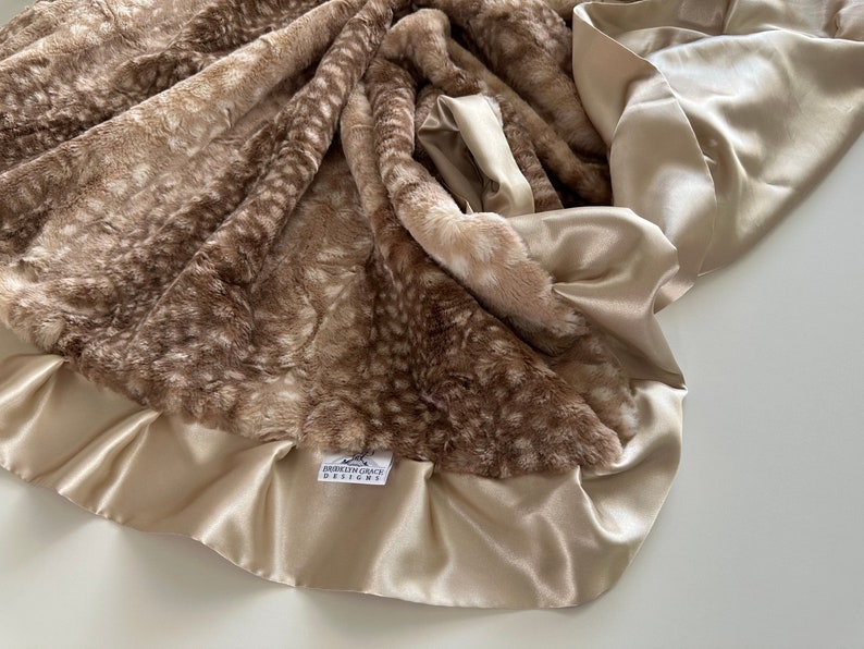 May include: A soft, brown and white faux fur baby blanket with a satin trim. The blanket has a subtle deer print pattern and a tag that reads "Everly Grace Designs".