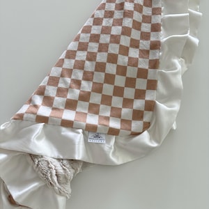May include: A baby blanket with a brown and white checkered pattern. The blanket has a white satin border with a ruffled edge. The blanket is soft and plush, with a tag that reads "BROOKLYN GRACE DESIGNS."