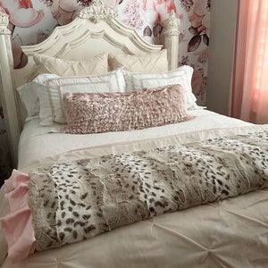 May include: A white bed frame with a tufted headboard and a white and pink bedding set. The bed is decorated with a pink throw pillow and a gray and white patterned throw blanket. The wall behind the bed is covered in a floral wallpaper with pink and white flowers.