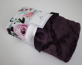 May include: A soft, plush blanket with a floral print on one side and a solid purple plush on the other side. The blanket is folded and tied with a purple ribbon.