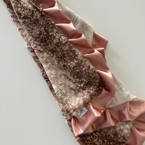 May include: A brown and beige faux fur baby blanket with a pink satin trim. The blanket has a ruffled edge and a small white tag that says "Precious Grace Designs".