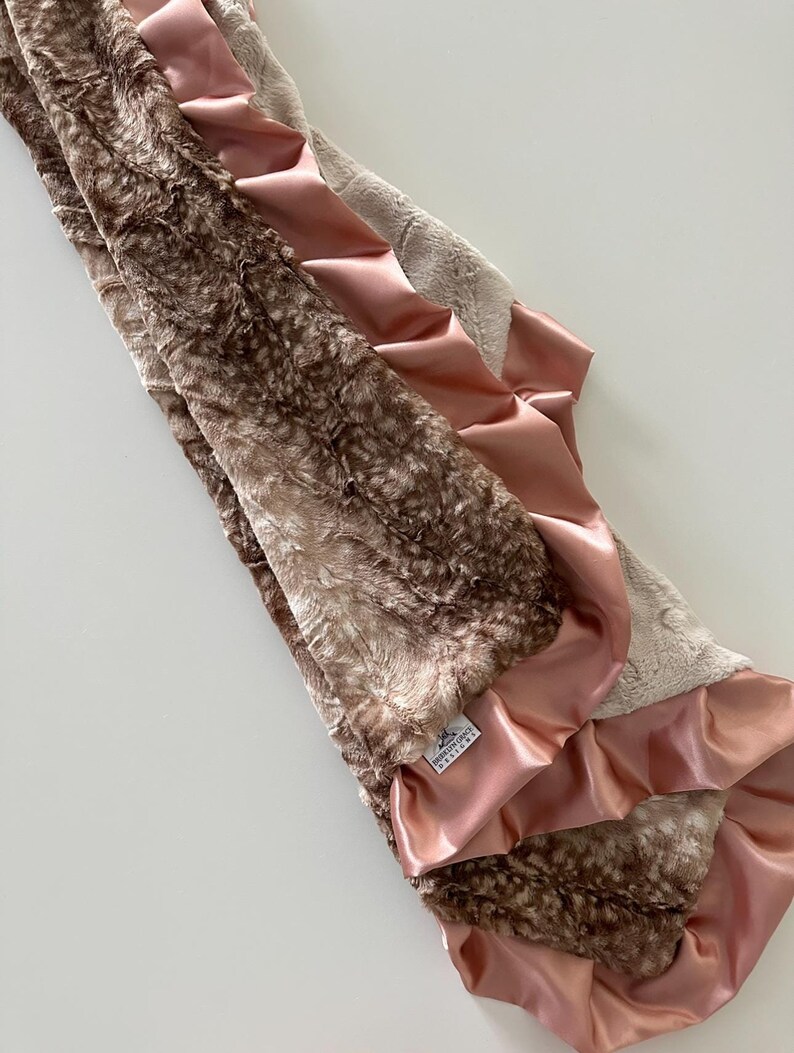 May include: A brown and beige faux fur baby blanket with a pink satin trim. The blanket has a ruffled edge and a small white tag that says "Precious Grace Designs".