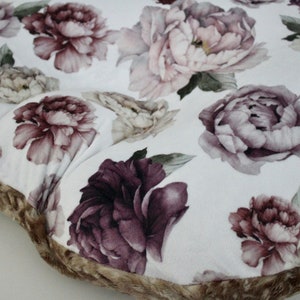 May include: A round, white cushion with a brown faux fur trim. The cushion is covered in a floral fabric with a pattern of pink, purple, and white peonies.