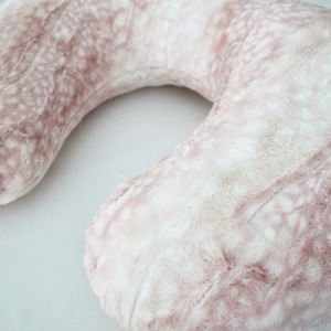May include: A pink and white faux fur travel neck pillow. The pillow is shaped like a horseshoe and has a soft, plush texture.