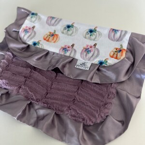 May include: A soft, purple and white baby blanket with a ruffled edge and a pumpkin print. The blanket has a tag that says "Birdsong Grace Designs".