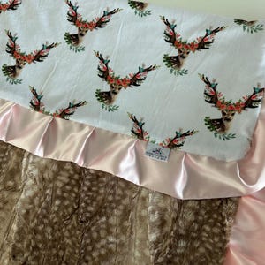 May include: A baby blanket with a white fabric top featuring a floral deer print and a pink satin trim. The bottom is a brown faux fur fabric.