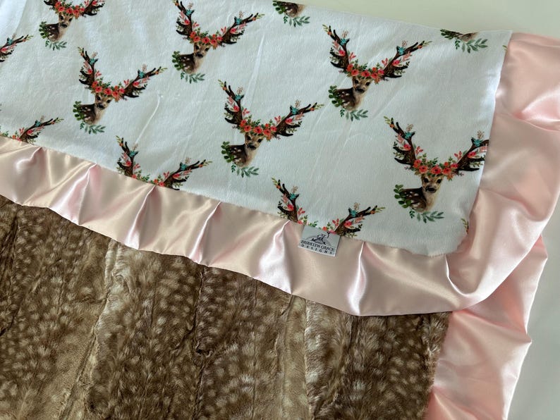 May include: A baby blanket with a white fabric top featuring a floral deer print and a pink satin trim. The bottom is a brown faux fur fabric.