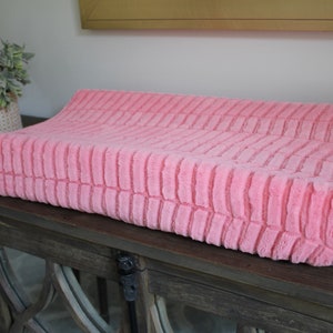 May include: A pink changing pad cover with a textured, ribbed design. The cover is made of soft, plush fabric.