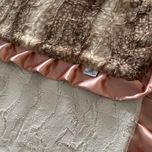 May include: A soft, plush baby blanket with a brown and white faux fur top layer, a pink satin backing, and a small white tag that reads "Brooklyn Grace Designs".