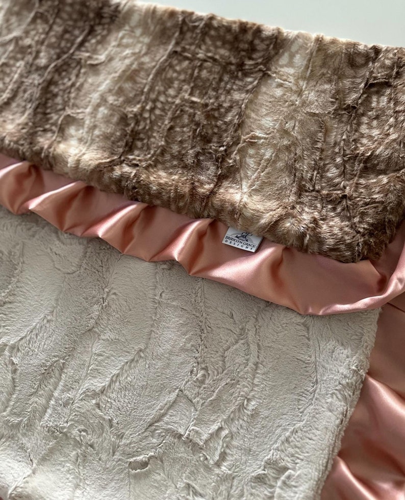 May include: A soft, plush baby blanket with a brown and white faux fur top layer, a pink satin backing, and a small white tag that reads "Brooklyn Grace Designs".