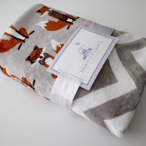 May include: A gray and white chevron patterned blanket with a gray and orange fox print on one side. The blanket is folded and has a white ribbon tied around it. A tag with the text "Brooklyn Grace Designs" is attached to the ribbon.