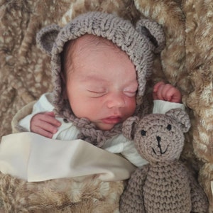 May include: A newborn baby wearing a gray knitted hat with bear ears and a white knitted onesie is sleeping on a brown faux fur blanket. A gray knitted teddy bear is next to the baby.