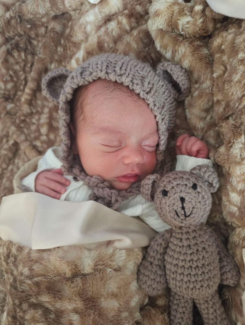 May include: A newborn baby wearing a gray knitted hat with bear ears and a white knitted onesie is sleeping on a brown faux fur blanket. A gray knitted teddy bear is next to the baby.