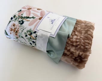May include: A rolled-up baby blanket with a floral pattern in pink, green, and brown. The blanket is tied with a sage green satin ribbon and a white ribbon. A tag with the text "Brooklyn Grace Designs" is attached.