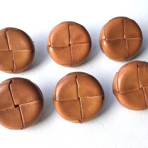 Puede incluir: Seis botones redondos de cuero marrón claro. Cada botón tiene un diseño cosido en cruz en el centro. Los botones están dispuestos sobre una superficie blanca. Adecuado para proyectos de costura y artesanía.