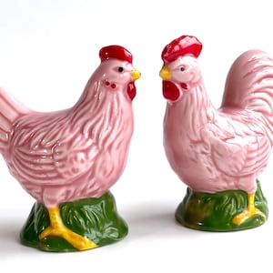 May include: Two pink ceramic chicken ornaments, a hen and a rooster. They have red combs and wattles, yellow beaks and feet, and sit on green bases. A delightful addition to a kitchen or dining room.
