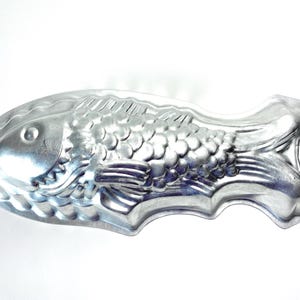 May include: A vintage, silver-colored fish-shaped baking mold. The mold has detailed scales, fins, and a handle. It is made of metal and is designed for baking cakes or other treats in the shape of a fish.