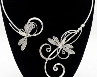 May include: A silver-toned wire choker necklace featuring two dragonfly motifs with intricate filigree wing patterns. The design incorporates swirling wire elements that extend from the dragonfly bodies, creating an open, asymmetrical collar structure.