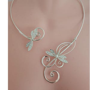 May include: A silver-toned wire collar necklace featuring two dragonfly motifs. The design includes intricate wire-wrapped details, swirling metal accents, and an open-front construction. The metallic finish is smooth and reflective throughout the piece.
