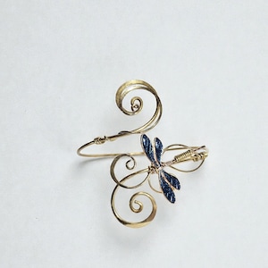 May include: A gold-toned bracelet with a stylized dragonfly design. The dragonfly has dark blue enamel wings and is centered on the bracelet. The bracelet features elegant, swirling metalwork.