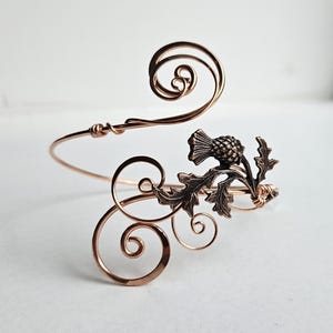 May include: A copper-colored arm cuff with a thistle design. The cuff features a swirling wire design with a detailed thistle and leaf accent. The metal has a warm, reddish-brown hue, and the design is intricate and delicate.