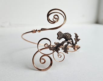 May include: A copper-colored arm cuff with a thistle design. The cuff features a swirling wire design with a detailed thistle and leaf accent. The metal has a warm, reddish-brown hue, and the design is intricate and delicate.
