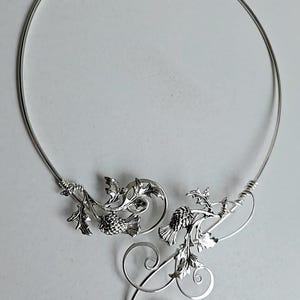 May include: A silver-toned necklace featuring a delicate thistle design. The necklace has a circular collar with detailed thistle and leaf embellishments. The design includes intricate swirls and a polished finish, suitable for special occasions.