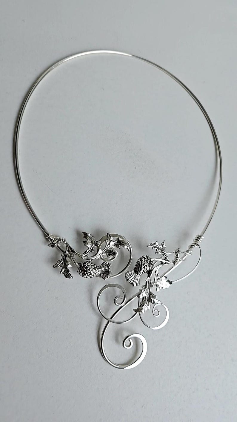 May include: A silver-toned necklace featuring a delicate thistle design. The necklace has a circular collar with detailed thistle and leaf embellishments. The design includes intricate swirls and a polished finish, suitable for special occasions.