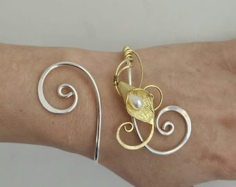 May include: A two-tone cuff bracelet featuring a silver-colored wire spiral on one side and a gold-colored wire design on the other. The gold section includes a textured calla lily flower shape with a single white pearl bead at its center.