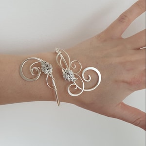 May include: Two silver-toned hair accessories. Each features a swirling design with a leaf-like accent. One has a long, pointed pin. The other has a curved design. The accessories are displayed on a light background.