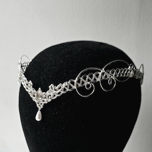 May include: A silver-colored tiara with an intricate wire design. The tiara features a central teardrop-shaped embellishment with a dangling pearl. The design includes woven patterns and delicate swirls, creating an elegant, ethereal look.