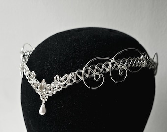 May include: A silver-colored tiara with an intricate wire design. The tiara features a central teardrop-shaped embellishment with a dangling pearl. The design includes woven patterns and delicate swirls, creating an elegant, ethereal look.