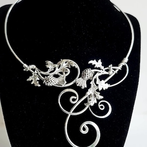 May include: A silver-toned necklace with a floral design, featuring leaves and pinecone-like elements. The necklace has a curved, open design with decorative swirls. It is displayed on a black mannequin.
