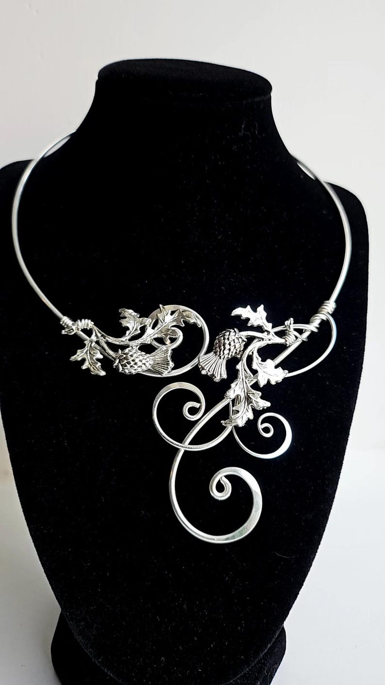 May include: A silver-toned necklace with a floral design, featuring leaves and pinecone-like elements. The necklace has a curved, open design with decorative swirls. It is displayed on a black mannequin.