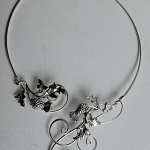 May include: A silver-toned necklace featuring a thistle design. The necklace has a circular shape with intricate floral and leaf details. The design includes spiraling accents and is set against a neutral background.