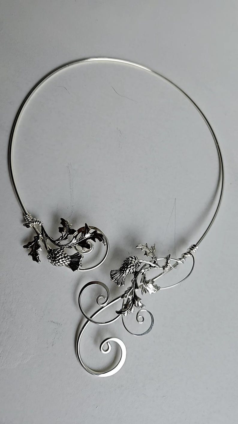 May include: A silver-toned necklace featuring a thistle design. The necklace has a circular shape with intricate floral and leaf details. The design includes spiraling accents and is set against a neutral background.