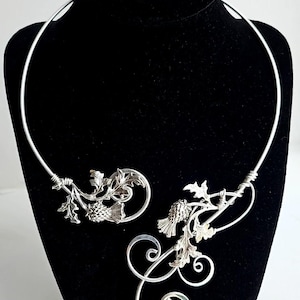 May include: A silver-toned necklace with a botanical design, featuring leaves and thistle-like elements. The necklace is displayed on a black mannequin, showcasing its intricate details and elegant form. The design includes curved lines and detailed metalwork.