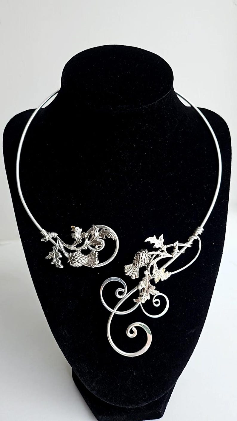 May include: A silver-toned necklace with a botanical design, featuring leaves and thistle-like elements. The necklace is displayed on a black mannequin, showcasing its intricate details and elegant form. The design includes curved lines and detailed metalwork.