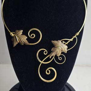 May include: A gold-toned metal collar necklace featuring an open front design. The necklace is adorned with two detailed leaf motifs and several wire-formed decorative swirls that extend from the ends of the rigid circular neck band.