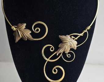 May include: A gold-toned metal collar necklace featuring an open front design. The necklace is adorned with two detailed leaf motifs and several wire-formed decorative swirls that extend from the ends of the rigid circular neck band.