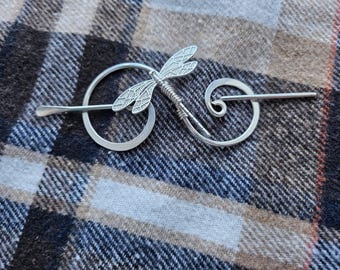 May include: A silver-colored hair pin with a dragonfly design. The pin features a dragonfly with detailed wings and a spiral design on the other end. The pin is resting on a plaid fabric.