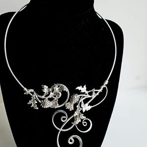 May include: A silver-toned necklace with a floral and scroll design. The necklace features detailed leaves, pinecone-like elements, and swirling accents. The piece is displayed on a black mannequin, highlighting its intricate craftsmanship.