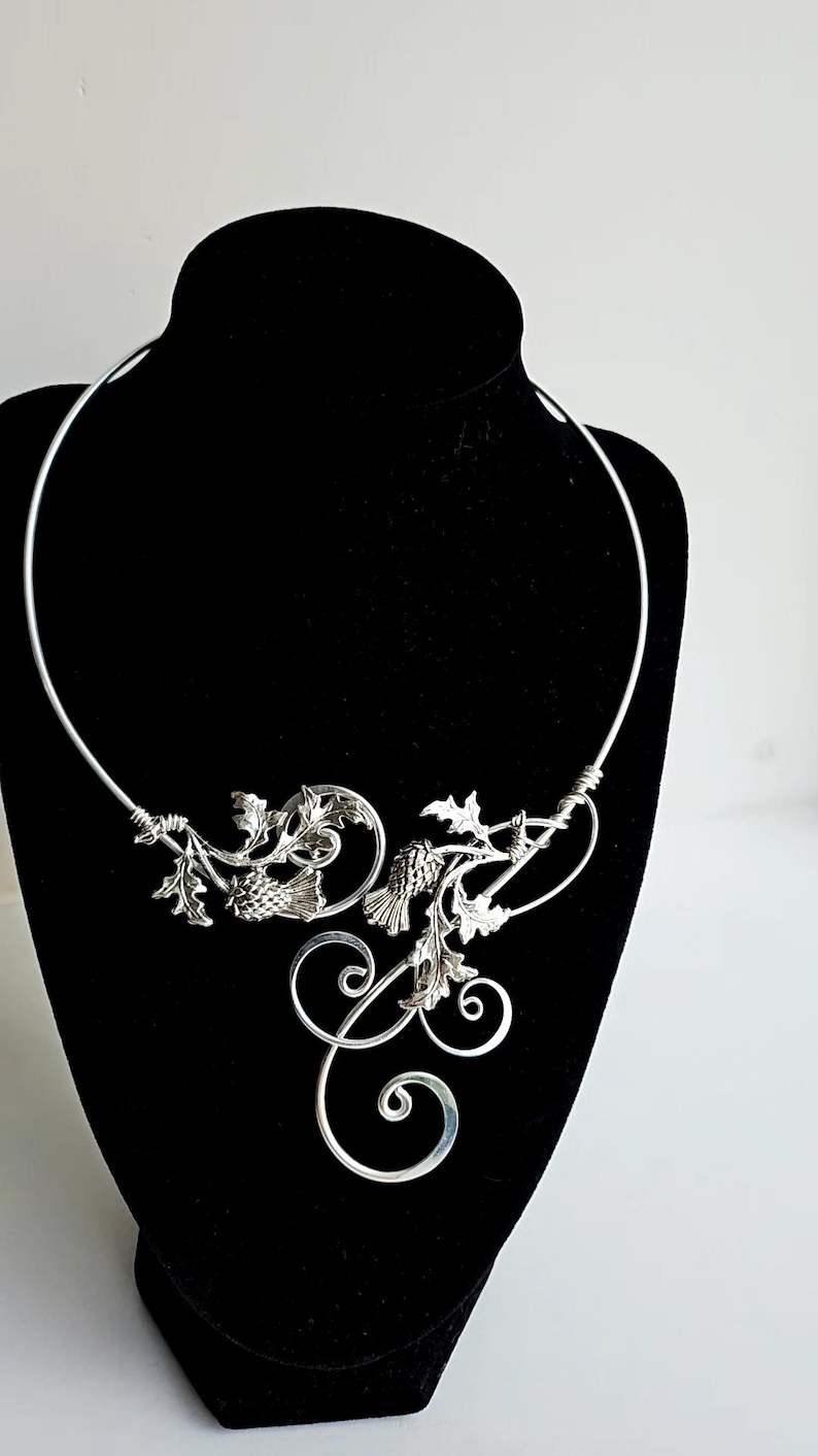 May include: A silver-toned necklace with a floral and scroll design. The necklace features detailed leaves, pinecone-like elements, and swirling accents. The piece is displayed on a black mannequin, highlighting its intricate craftsmanship.