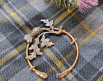 May include: A circular copper brooch featuring a detailed thistle motif with textured leaves and a flower head. The metal has a warm, burnished finish, and the design is secured with a wrapped wire detail, creating an open, hoop-like structure.